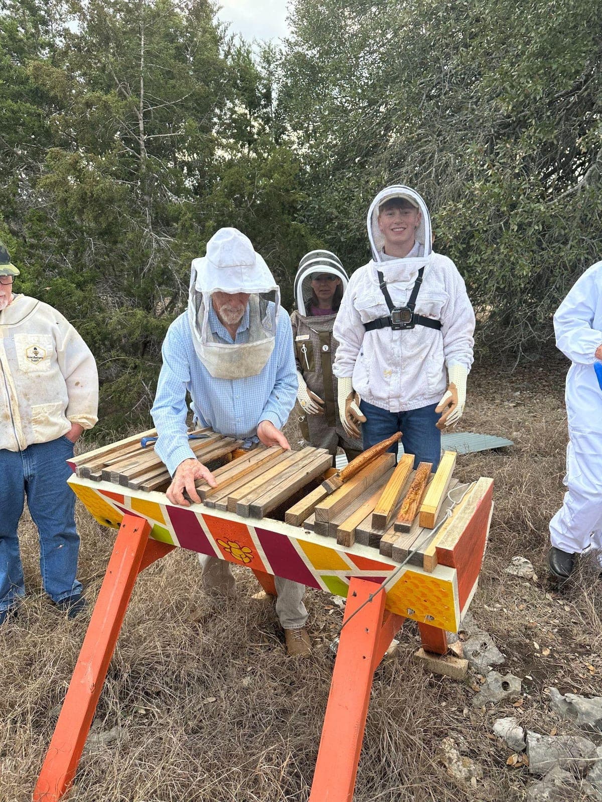 Joshua Davis in a bee suit and veil at a beekeeping workshop, standing beside an older beekeeper working the bars of a brightly painted top-bar hive