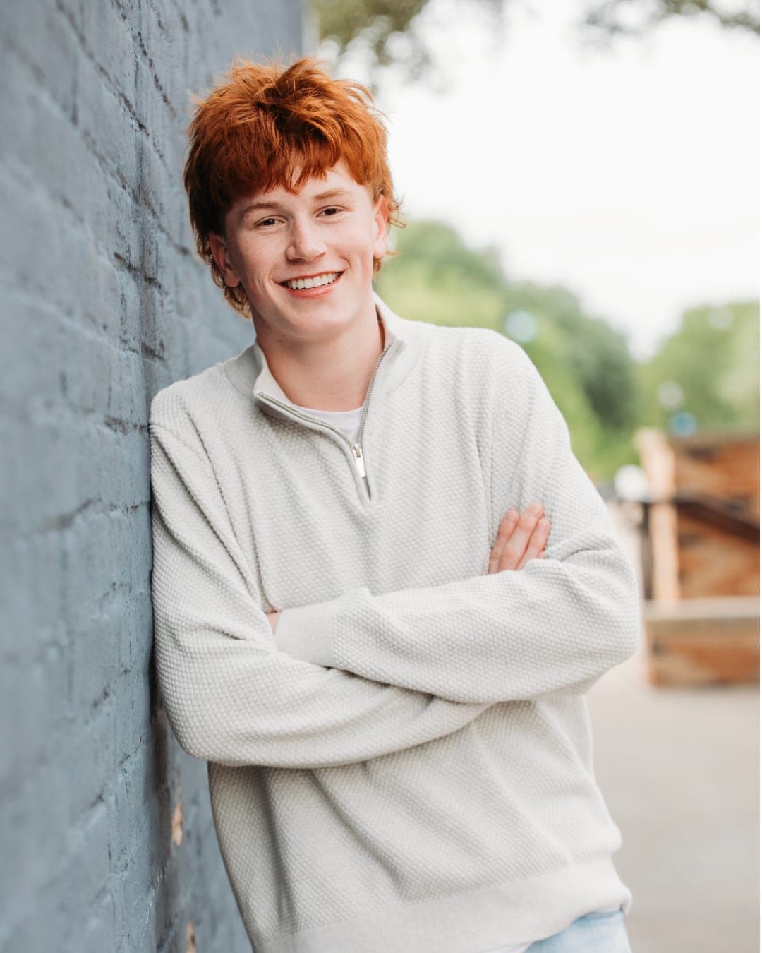 Joshua Davis, arms folded, leaning against a painted brick wall in a cream quarter-zip sweater