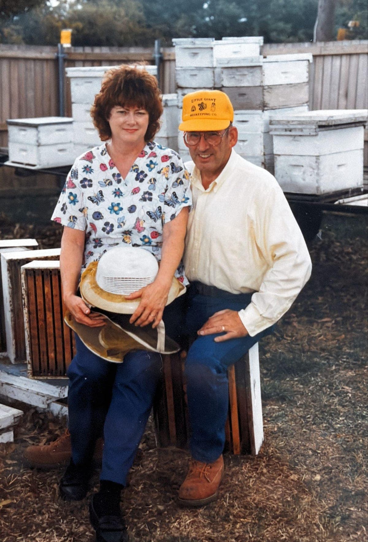 Louise and Harold, who founded the business in 1980, seated among stacked white hive boxes; Harold wears a Little Giant Beekeeping Co. cap